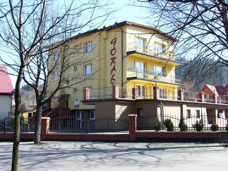 Żółty, trzypiętrowy budynek z balkonami i napisem 'GÓRAL' na elewacji, otoczony niskim, ceglanym murem z metalowym ogrodzeniem, Rajcza.